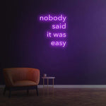 Elegant purple 'Nobody Said It Was Easy' LED neon sign, a stylish and motivational decor piece for your office, gym, or bedroom, reminding you of the power of perseverance.